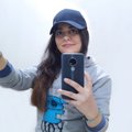 A small, low-resolution photo of a woman with long dark hair wearing a blue cap and gray sweatshirt, taking a mirror selfie with her phone.