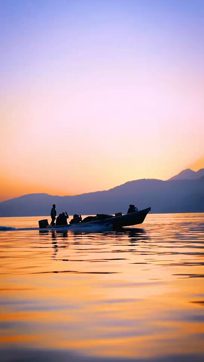 The image shows a boat sailing across a body of water at sunset with mountains in the background. There are two people visible on the boat, one person standing near the front and another sitting further back. The scene captures the beauty of nature as the sun sets over the horizon, creating a serene atmosphere for those aboard the boat.