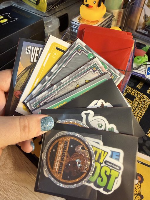 A hand with glittery teal nails holding a fan of Pokemon trading cards, including a visible "Pikachu" themed coin card, on a desk cluttered with card accessories.