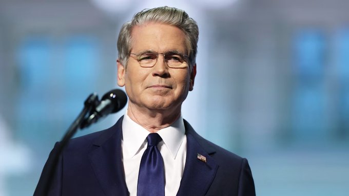 Treasury Secretary Scott Bessent stands at a podium in a dark blue suit with an American flag lapel pin, as the post references a $61 million crypto seizure tied to Iranian oil sales laundered through Binance. His official appearance lends authority to the U.S. Treasury's announcement of the civil forfeiture action against Chinese companies.