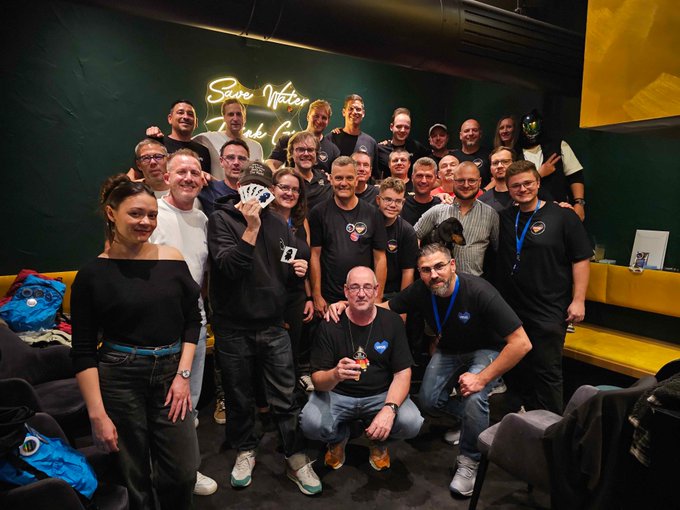 Group photo of approximately 25 people gathered in front of a neon sign reading 'Save Water Drink [Beer]', with one person holding up a large card.