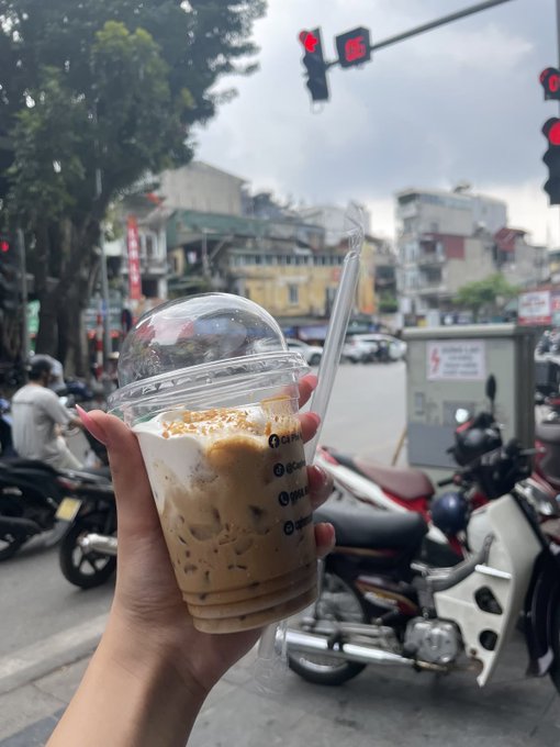 A hand holding a plastic cup of iced coffee with a straw, taken on a street with motorbikes and traffic lights in the background.