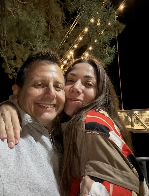 A selfie of a smiling man and woman at night, with the woman wearing a brown and red patterned jacket, set against a backdrop of string lights and trees.