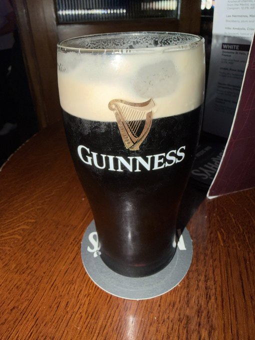 A pint glass of Guinness beer with its signature harp logo sits on a coaster on a wooden table.