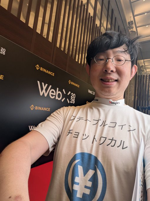 A selfie of a man wearing glasses and a white t-shirt with Japanese text and a yen symbol, standing in front of a backdrop with "BINANCE" and "Web3 2026" logos.