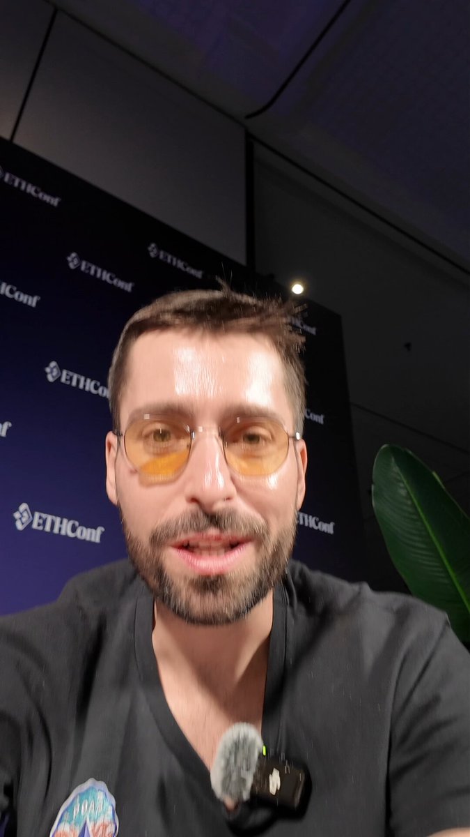The image features a man wearing glasses and a black shirt who appears to be smiling while taking a selfie. The background of the photo includes a banner that says "Ethiccon" and "EITC". There are also some text elements, such as "ethiccon" and "EITC", which might suggest that this is related to an event or conference.