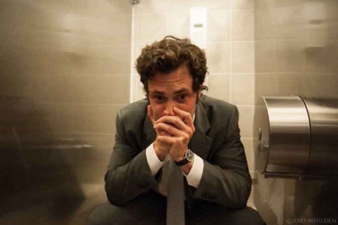 A man in a gray suit sits on a toilet in a bathroom stall, covering his mouth with his hands in distress.