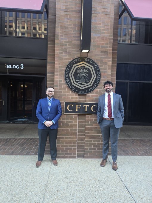 Two men in business suits stand smiling in front of the brick building entrance marked "CFTC" with the "Commodity Futures Trading Commission" seal.