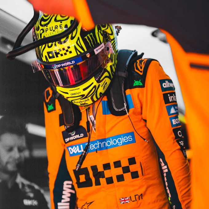A McLaren Formula 1 driver in an orange racing suit and yellow helmet prepares in the garage, with visible sponsors including Dell Technologies, OKX, DP World, and a British flag marked "LM" on the chest. The image embodies the post's message about year-round dedication, showing the team grinding through summer preparation to keep building toward performance.