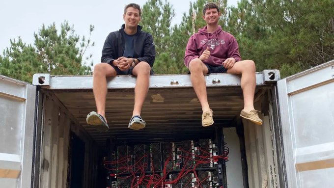 Two young men sitting on the edge of an open shipping container filled with computer servers and red cables, surrounded by trees.