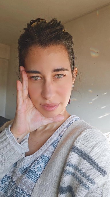 A woman with her hair in a messy bun poses for a selfie, resting her hand against her cheek while wearing a striped knit sweater, sharing a soft "Good Morning" greeting with her followers.