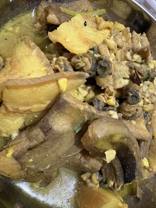 Close-up of a stewed dish featuring pork belly and what appears to be chicken or duck in a yellowish broth with herbs and garlic in a pot.