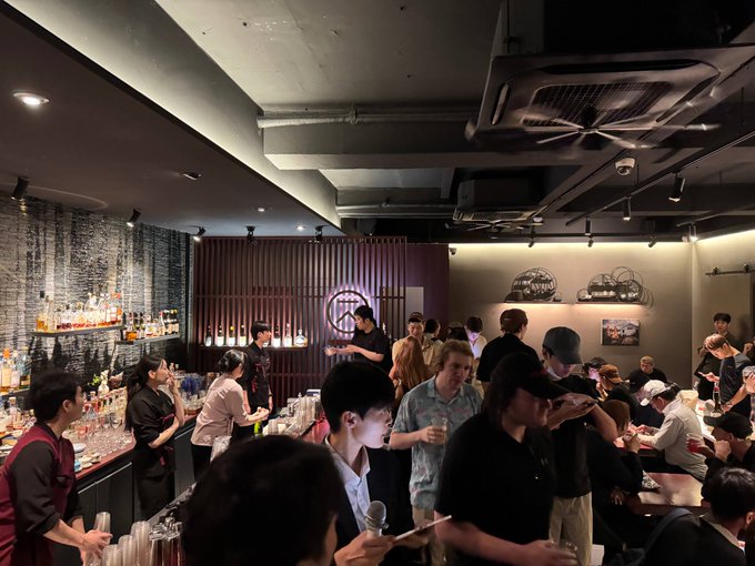 A crowded indoor venue, possibly a bar or restaurant, with people gathered around a counter where bartenders in black and red uniforms are preparing drinks.