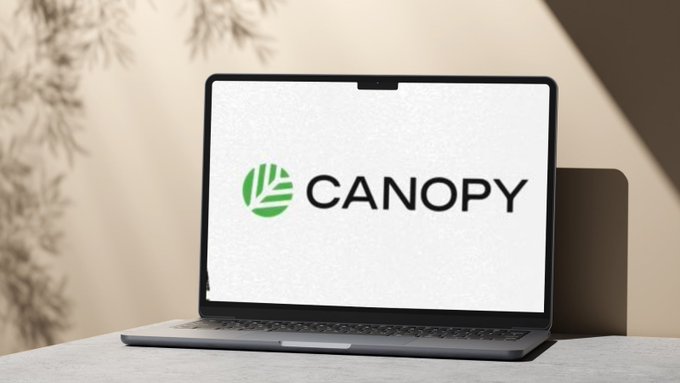 A laptop on a desk displaying the green and white 'CANOPY' logo, with a plant visible in the softly lit background.