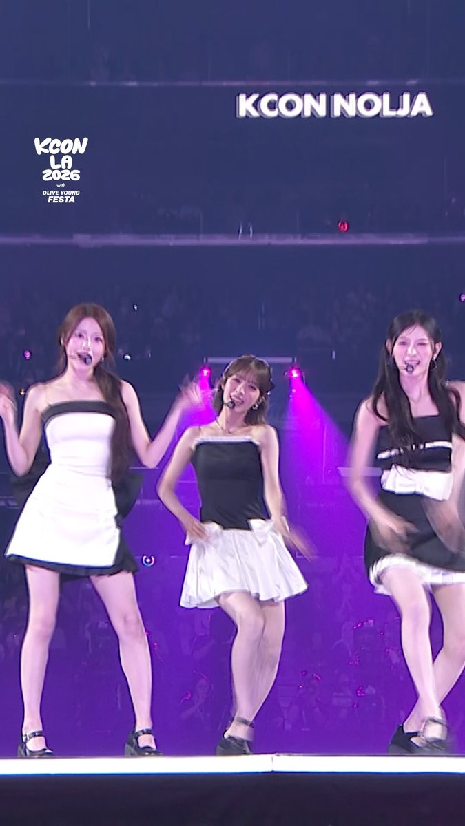 Three female K-pop performers from the group RESCENE perform on stage at KCON LA 2026, dressed in coordinated black and white outfits with short skirts, while on-screen graphics display "KCON NOLJA" and "KCON LA 2026 with OLIVE YOUNG FESTA" during their rendition of "LOVE ATTACK" on M Countdown's Stage Cam on Day 3.
