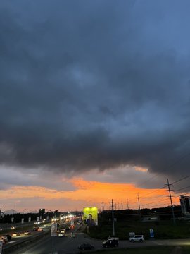 A view of a sunset with dark storm clouds over a road with traffic, power lines, and industrial structures in the distance.