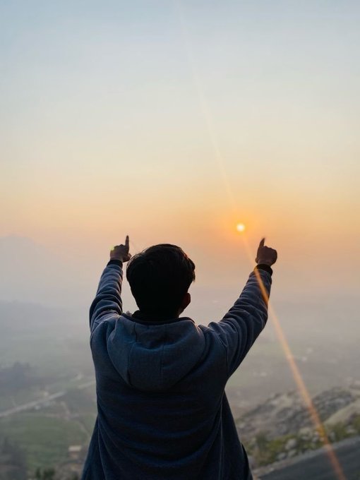 A person viewed from behind, wearing a blue hoodie, pointing both index fingers toward the setting sun over a hazy mountain landscape.