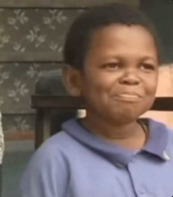The image shows a young boy wearing a blue shirt with his head tilted back as if he's looking at something or someone off camera. The background features a chair and a wall with a floral pattern, creating an indoor setting for the scene.