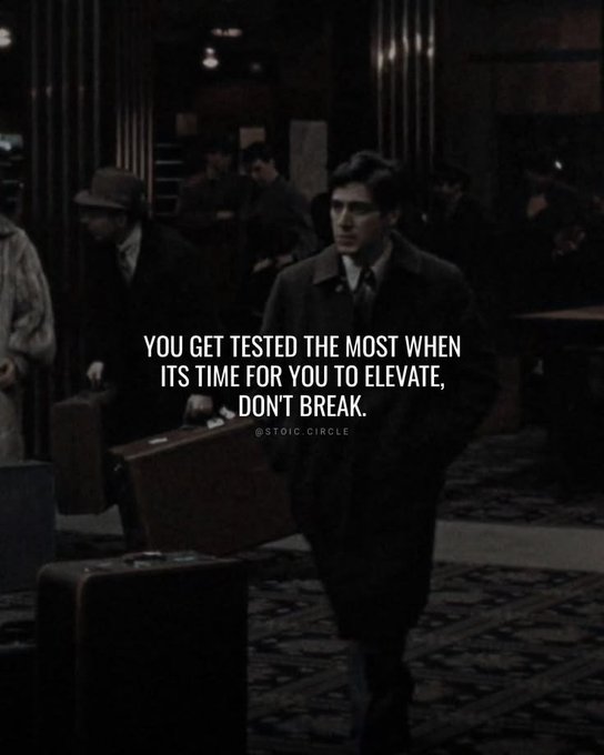 A dark, cinematic image of a man in a trench coat carrying a suitcase, overlaid with the motivational text: 'YOU GET TESTED THE MOST WHEN ITS TIME FOR YOU TO ELEVATE, DON'T BREAK.' - @stoic_circle