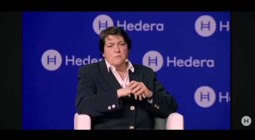 The image shows a woman sitting in an office chair, wearing a suit jacket with a pinstripe pattern. She appears to be giving a speech or presentation, as indicated by her body language and the fact that she has her hands together on her lap. The background of the image is blue, which could potentially represent a company logo or color scheme for the event where the woman is speaking.