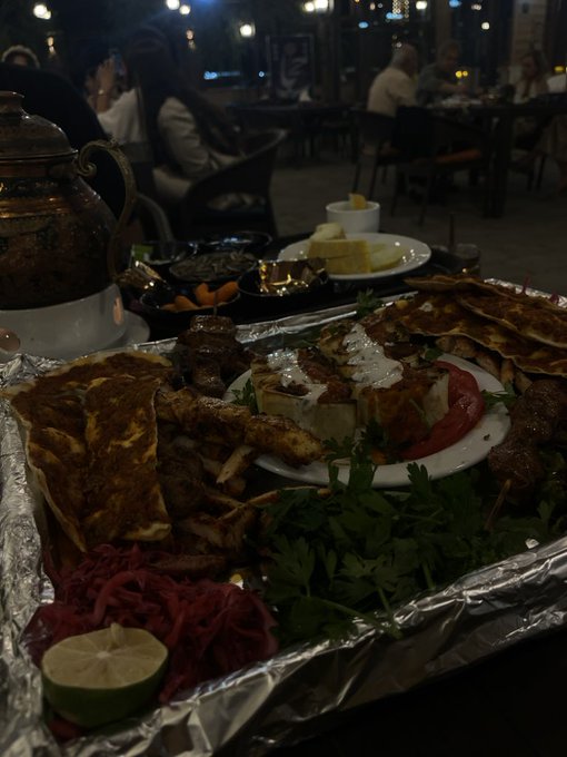 A dimly lit restaurant table is laden with a feast of Middle Eastern cuisine, including flatbread, grilled meats, fresh parsley, sliced tomatoes, pickled red cabbage, and a lime half, with a copper teapot nearby. The spread pairs perfectly with "Gn bros," suggesting the meal is being shared among good friends enjoying a hearty, shared dining experience.