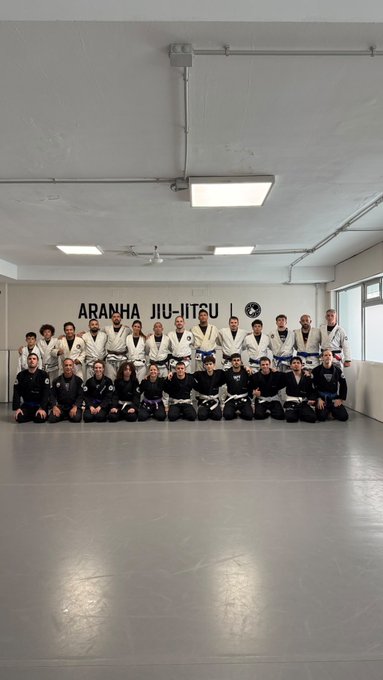 A group photo of approximately 24 martial arts practitioners in gis, some wearing white and others black, posing together on mats in a training hall with the wall text 'ARANHA JIU-JITSU' behind them.