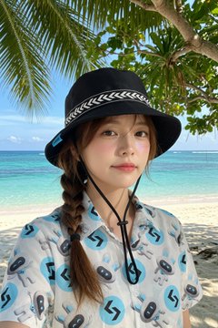 A young woman wearing a black bucket hat and a patterned shirt taking a selfie at a tropical beach with palm trees and turquoise water.