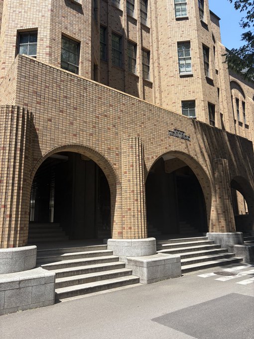 A photograph of a large brick building with three prominent arched entryways and stone steps leading up to them, bathed in bright sunlight.