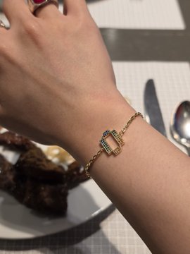 A close-up photo of a person's wrist wearing a delicate gold bracelet with a rectangular green gemstone centerpiece, taken over a dining table setting.