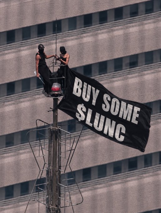 Two protesters standing on a tall antenna or tower structure, holding a black banner that reads 'BUY SOME $LUNC', with a large office building featuring many windows in the background.