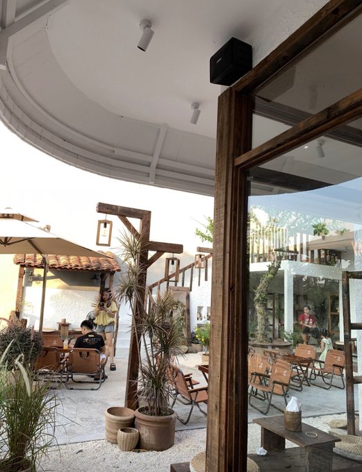 An outdoor courtyard cafe area with wooden tables and chairs, potted plants, and people sitting under a tiled roof overhang on a sunny day.