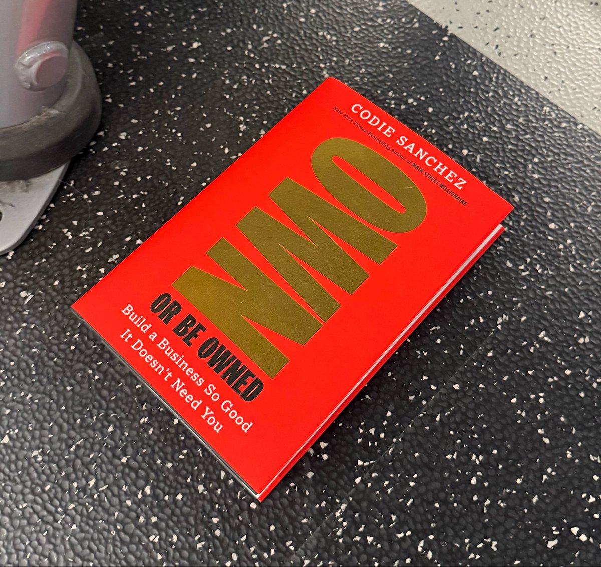 A red book titled 'WHO NOT HOW OR BE OWNED: Build a Business So Good It Doesn't Need You' by Codie Sanchez sits on a black textured surface next to gym equipment.