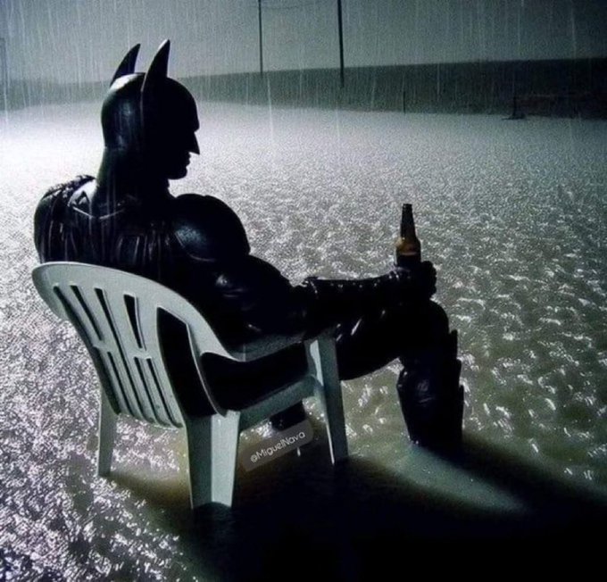 Batman sits in a white plastic lawn chair in the pouring rain, casually holding a bottle, in a moody, cinematic still.