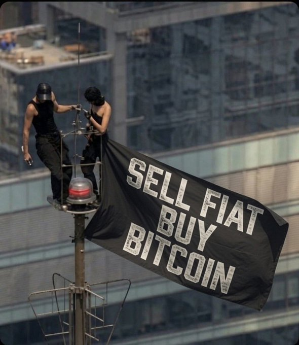 Two people stand on a rooftop antenna platform unfurling a black banner reading 'SELL FIAT BUY BITCOIN' against a city skyline.