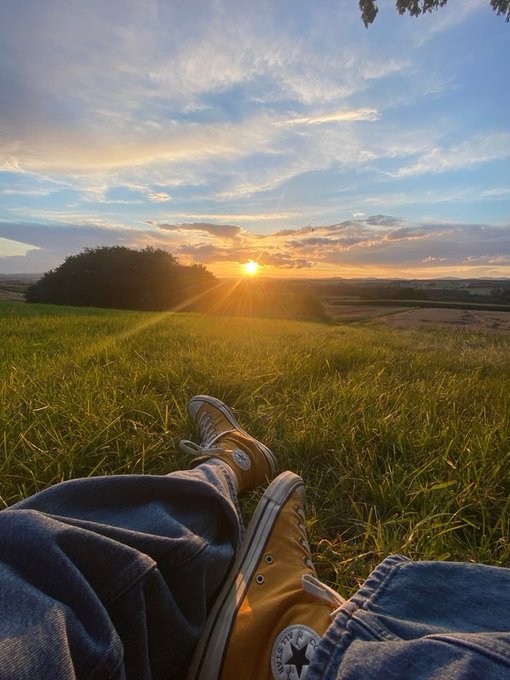 A first-person perspective shows someone relaxing in a grassy field at sunset, wearing jeans and yellow Converse sneakers, looking out over a rural landscape.