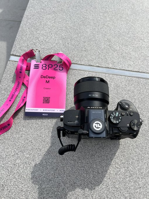 A Sony camera with a lens and attached Rode microphone lying on concrete next to a pink 'BP25' DeDeep M Creator badge from BREAKPOINT.