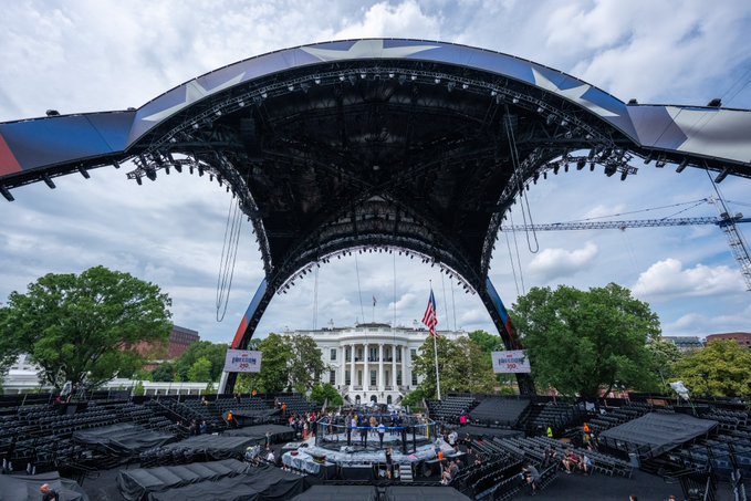 The image shows a large outdoor event taking place at the White House, with a stage set up for an event featuring UFC fighter #UFCWhiteHouse. The stage has a unique design resembling a tent or a canopy, providing shade and protection from the elements. There are several people gathered around the stage, possibly attending to various tasks related to the event. In addition to the main stage, there is also a smaller stage in the background with a flag on it.