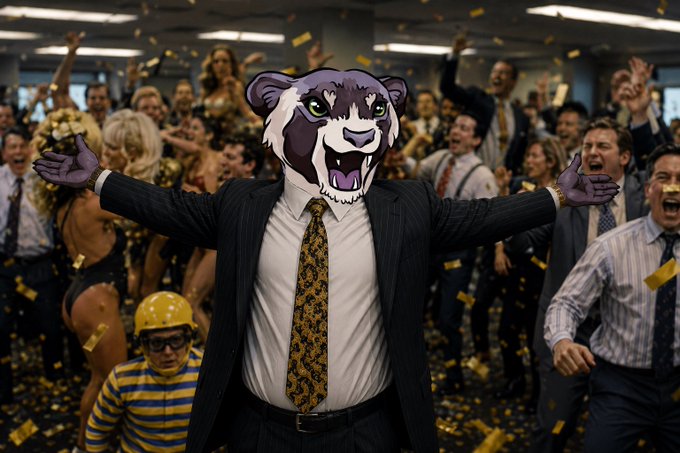 A figure in a pinstripe suit with a cartoonish purple ferret/wolverine mask stands triumphantly with arms outstretched amid a chaotic office celebration, as confetti rains down and cheering coworkers—including a hard-hatted construction worker—surround the scene. The surreal mascot imagery and defiant "We're not fucking leaving" post tie directly to the $ferret ticker, evoking a meme-worthy rally crowd refusing to sell.
