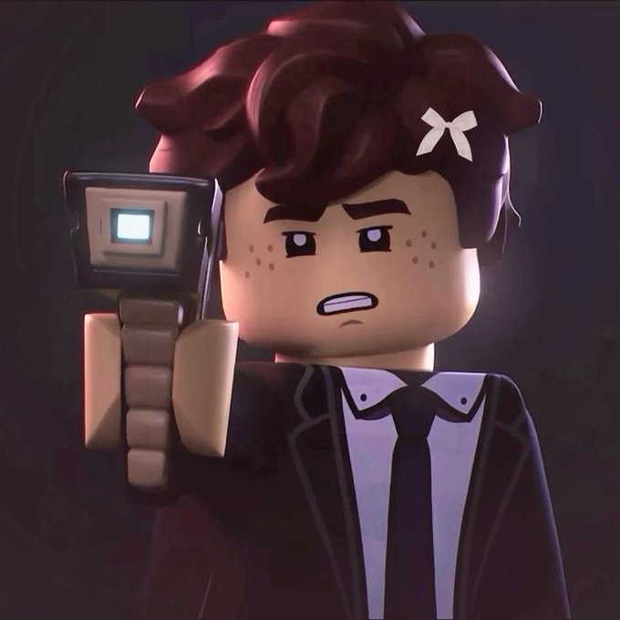 A 3D animated LEGO-style character in a black suit and tie with a white bow in their hair holds up a device with a glowing blue screen.