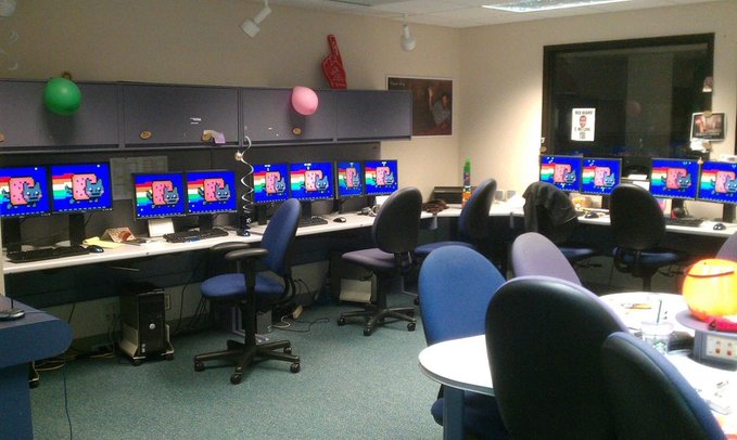 A computer lab with multiple monitors displaying identical blue screens, rows of office chairs, and decorations like green and pink balloons hanging from the ceiling.