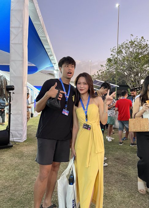 A man and a woman posing together at an outdoor event with blue tents in the background; both are wearing lanyards and making hand gestures, with the woman in a yellow dress holding a tote bag.