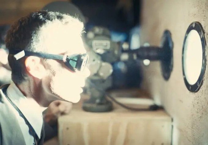 A man wearing safety goggles looks through a porthole or lens in a dimly lit industrial or laboratory setting.