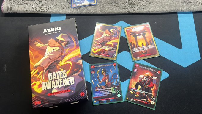 An Azuki Trading Card Game 'Gates Awakened' Starter Deck box displayed alongside four character cards on a blue playmat.