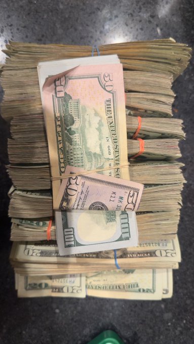 A stack of bundled U.S. $50 and $20 bills sits on a dark countertop, with one $50 bill featuring a gold-foil reverse flipped over the top. The post jokingly suggests airdropping the cash to $ANSEM token holders, humorously framing the physical money as a crypto giveaway.