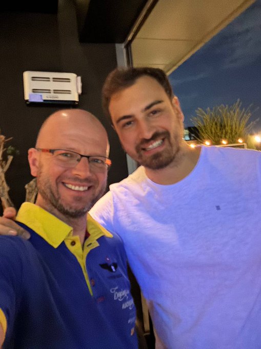 Two smiling men pose for a selfie at night; the man on the left has glasses and wears a blue and yellow shirt, while the taller man on the right wears a light blue shirt.