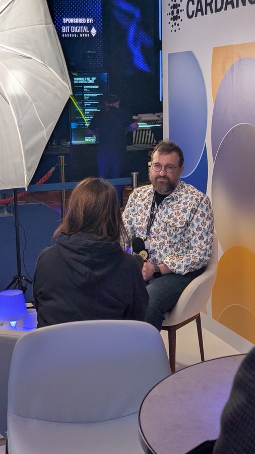 A bearded man with glasses in a patterned shirt is being interviewed at a Cardano-sponsored event with 'BIT DIGITAL' signage visible in the background.