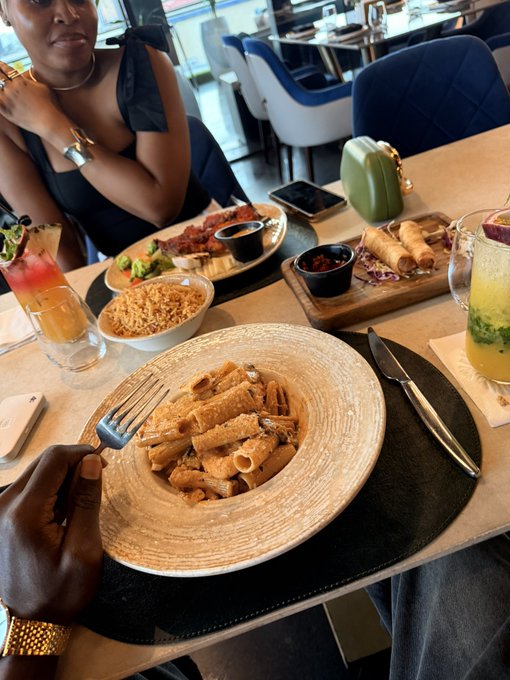 A restaurant table with a bowl of rigatoni pasta, a glass of yellow drink, rice, and appetizers, with a person sitting across the table.