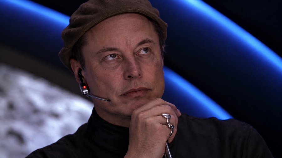 A close-up portrait of Elon Musk wearing a brown beanie and black turtleneck, with a headset microphone near his face and his hand resting near his chin.