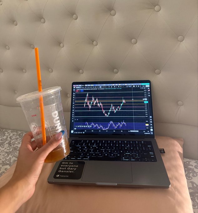 A person's hand holds a partially empty plastic Dunkin' cup with an orange straw next to a laptop displaying a cryptocurrency trading chart, with a laptop sticker reading 'GM to everyone but Gary Gensler.'