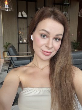 A selfie of a woman with long brown hair wearing a white strapless top and a gold necklace, posing in a lounge or lobby area with white sofas.